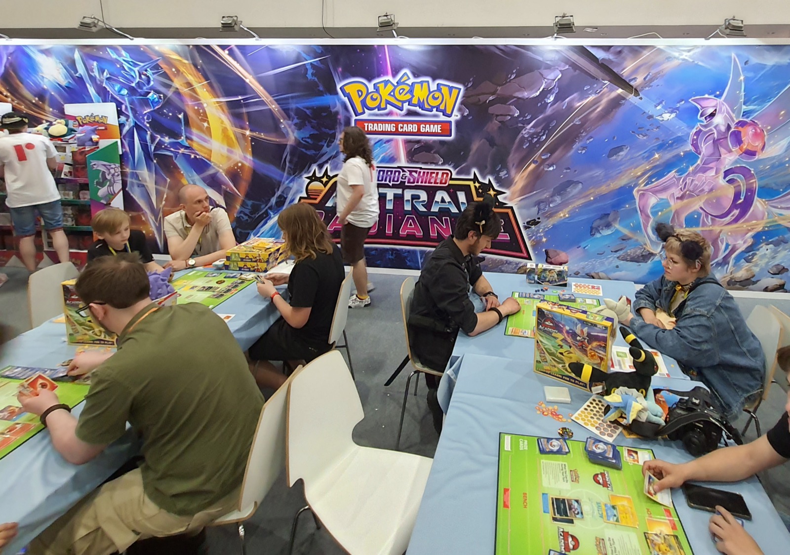 stand d'un tournoi Pokémon Trading Card Game avec des joueurs assis autour de tables, cartes et boosters posés devant eux
