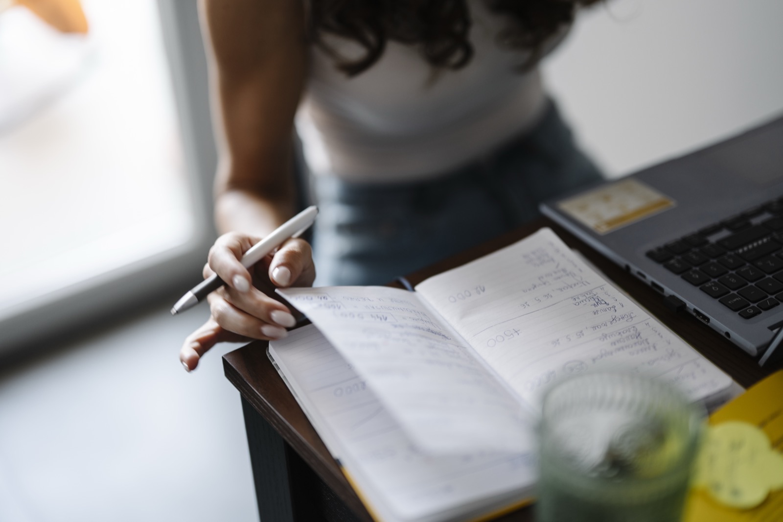 main qui écrit dans un carnet de notes ouvert, stylo à la main, devant un ordinateur portable